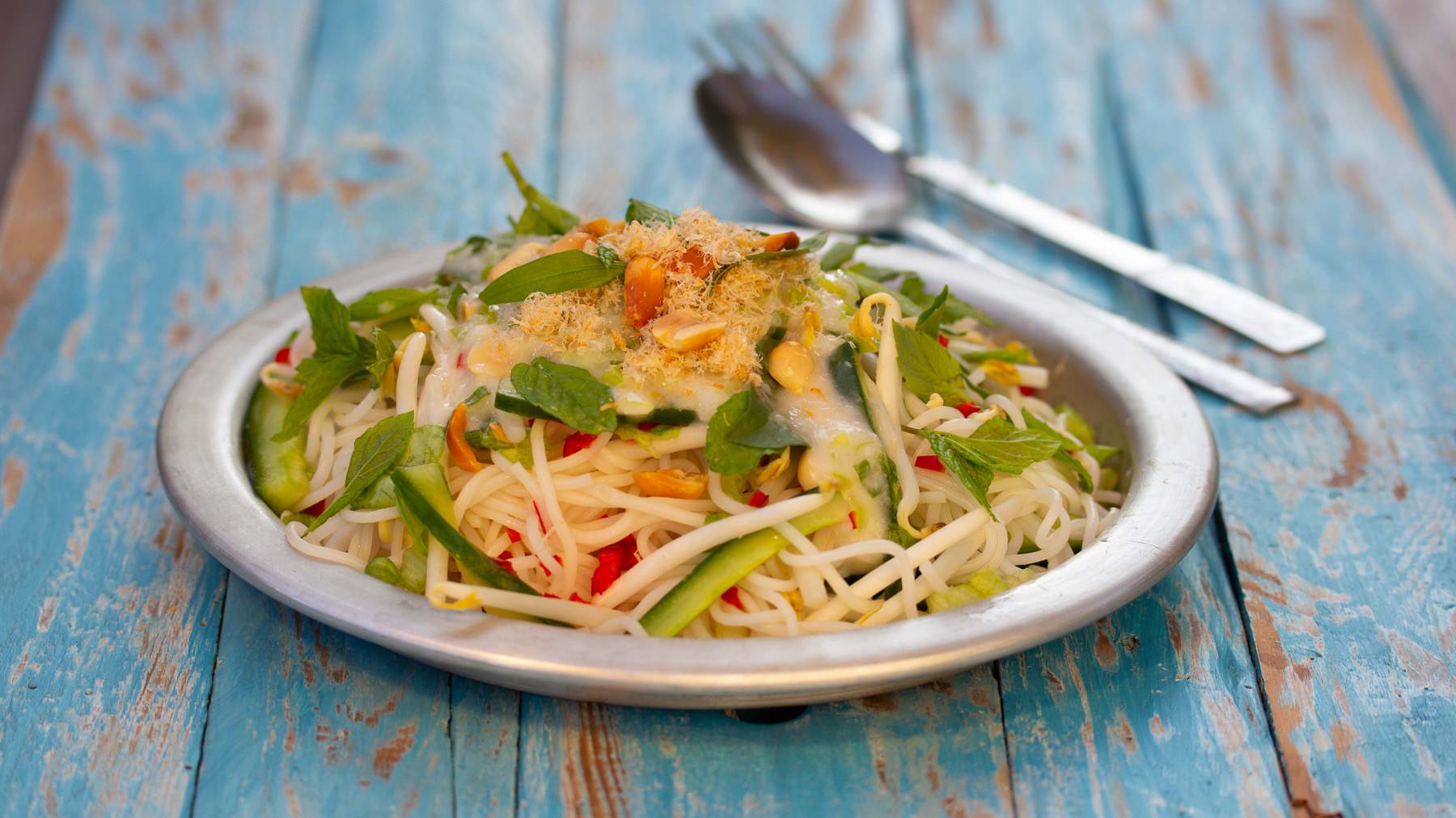 Reisnudelsalat mit Kokosdressing aus Kampot: Mitten im Gewusel des Morgenmarkts von Kampot entdecken wir eine Marktfrau mit einem Bambusjoch über den Schultern, zwei schwere Körbe im Gleichgewicht. Sie hält zwischen Fischständen an, klappt ein...